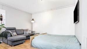 Een moderne, minimalistische kamer met een grijze bank, een klein tafeltje, een lamp en een bed met een blauwe deken.