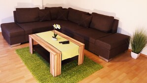 A modern living room with a brown sectional sofa, a wooden coffee table with a small plant, and a painting of a forest on the wall. - Gräfrath Gästehaus Neunkirchen-Seelscheid (Neunkirchen-Seelscheid)
