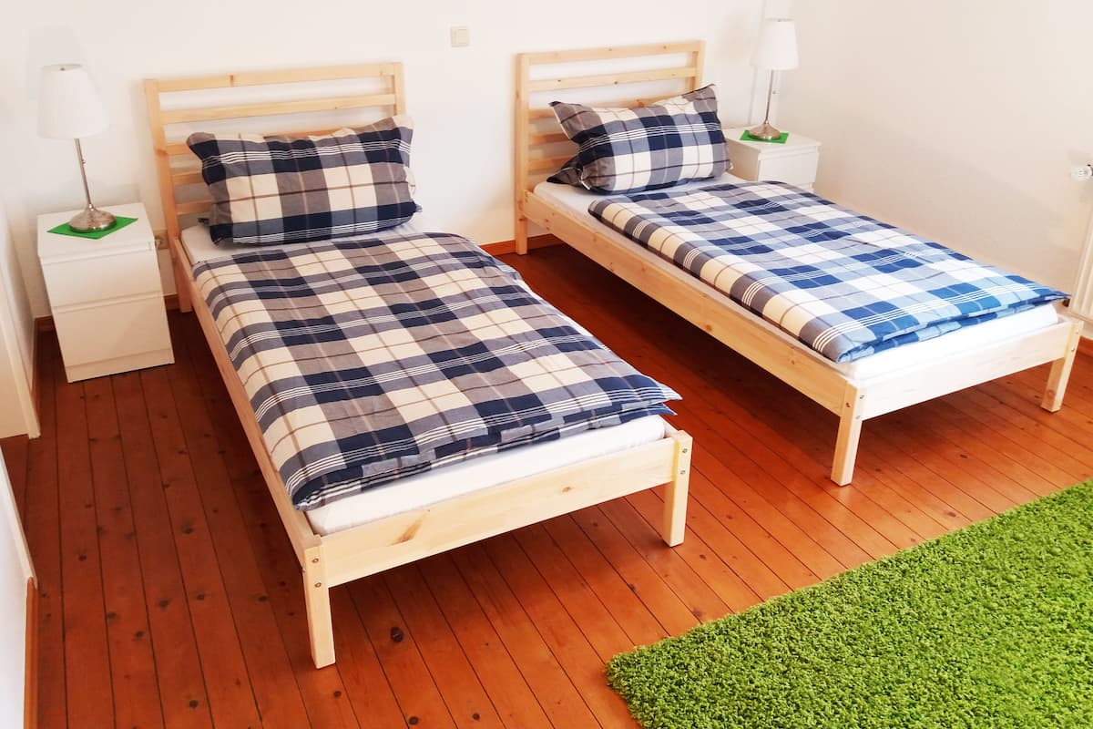 Two wooden beds with checkered bedding, white nightstands with lamps, and a green rug on a wooden floor.