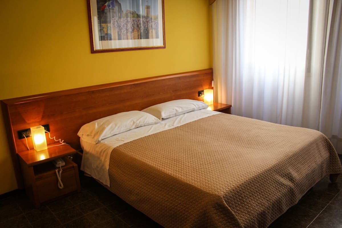 A hotel room with a wooden headboard, a bed with a brown quilt, two bedside lamps, a nightstand with a telephone, and a framed picture on the wall.