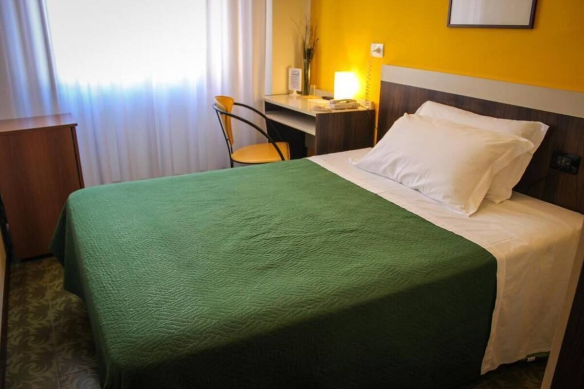 A hotel room with a green bedspread, a wooden desk, a chair, and a bedside lamp.