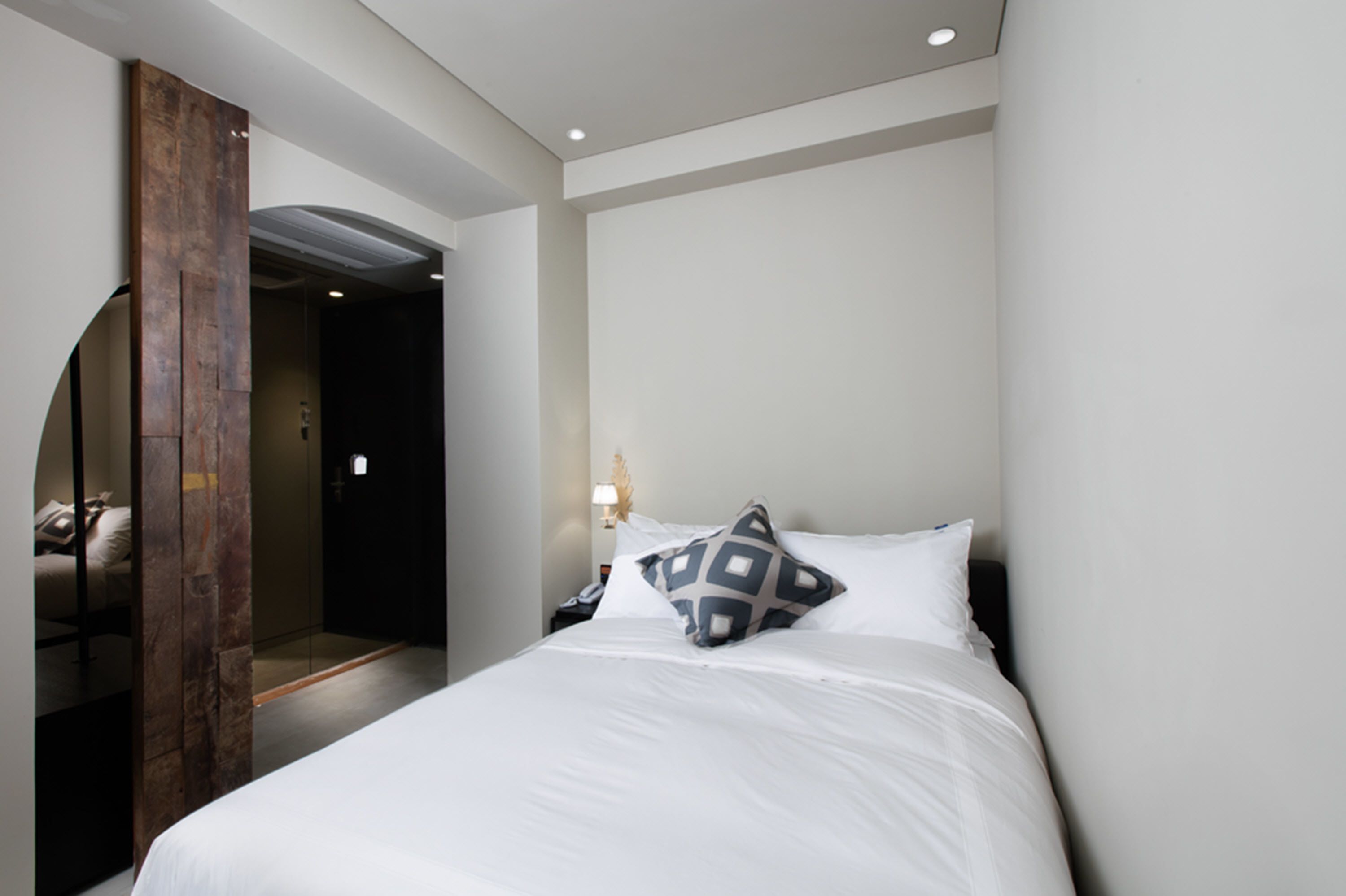 A hotel room with a bed, a wooden door, a nightstand, and a wall-mounted lamp.