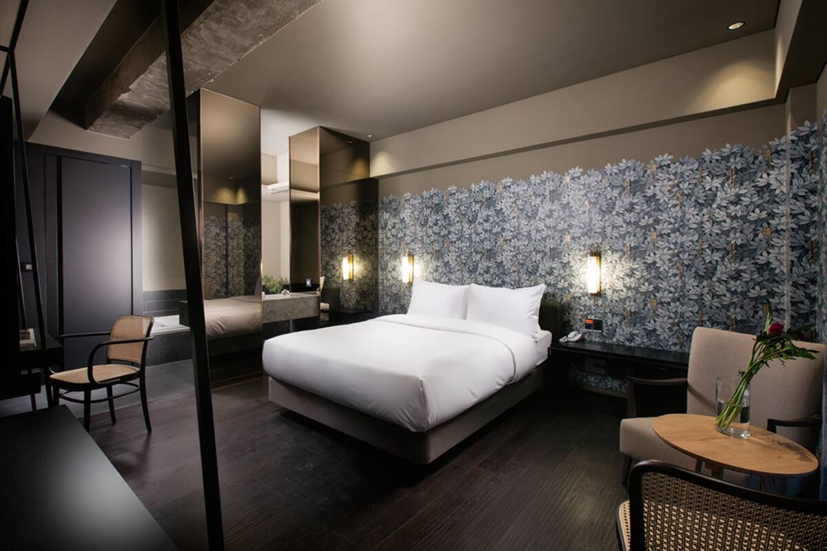 A modern hotel room with a large bed, a sitting area with a table and chairs, and a decorative wall with a floral pattern.