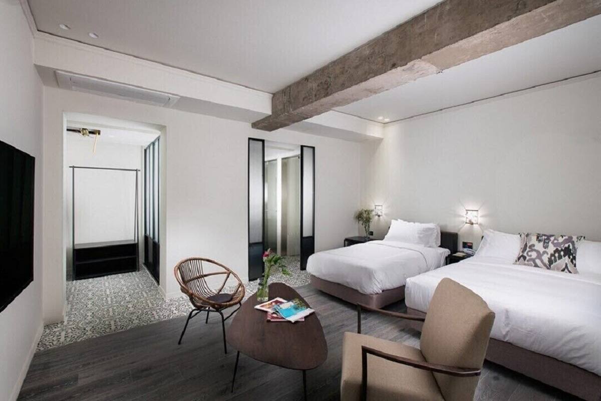 A modern hotel room with two beds, a sitting area with a chair, a small table, and a large mirror.