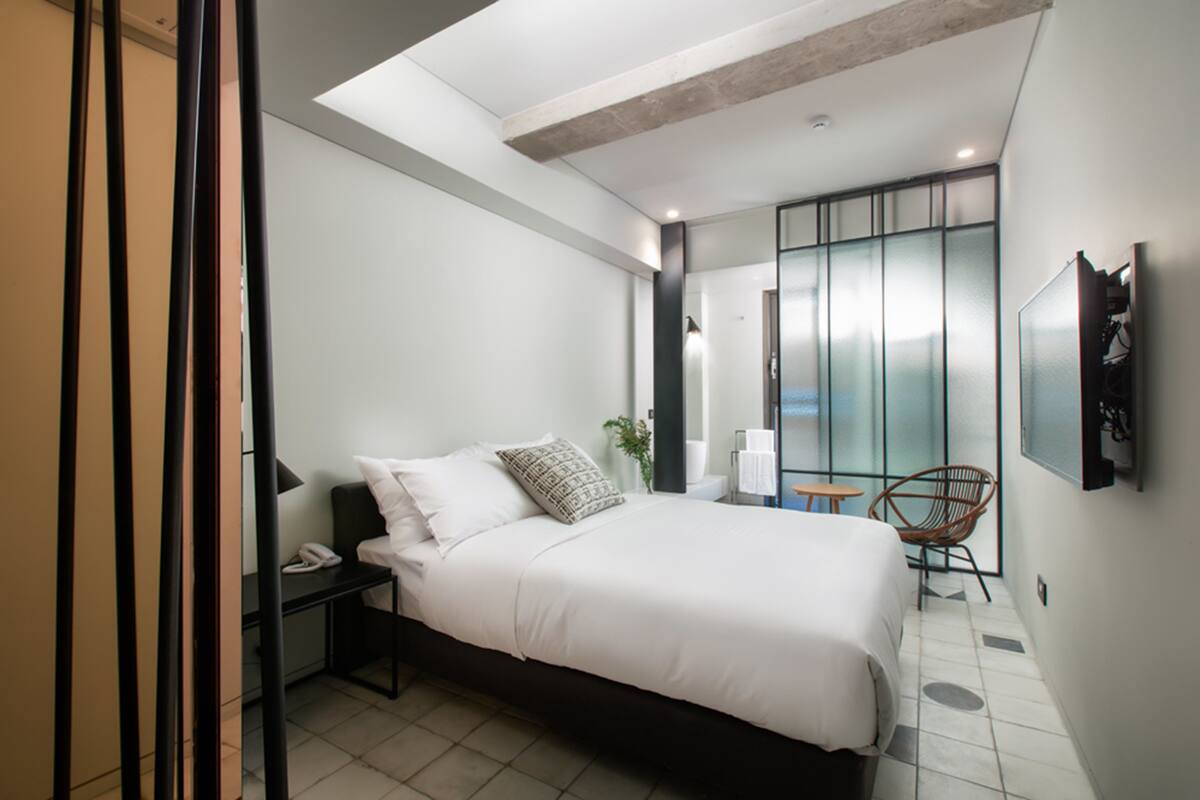 A modern bedroom with a large bed, a bedside table with a phone, a chair, and a view of the city through a large window.