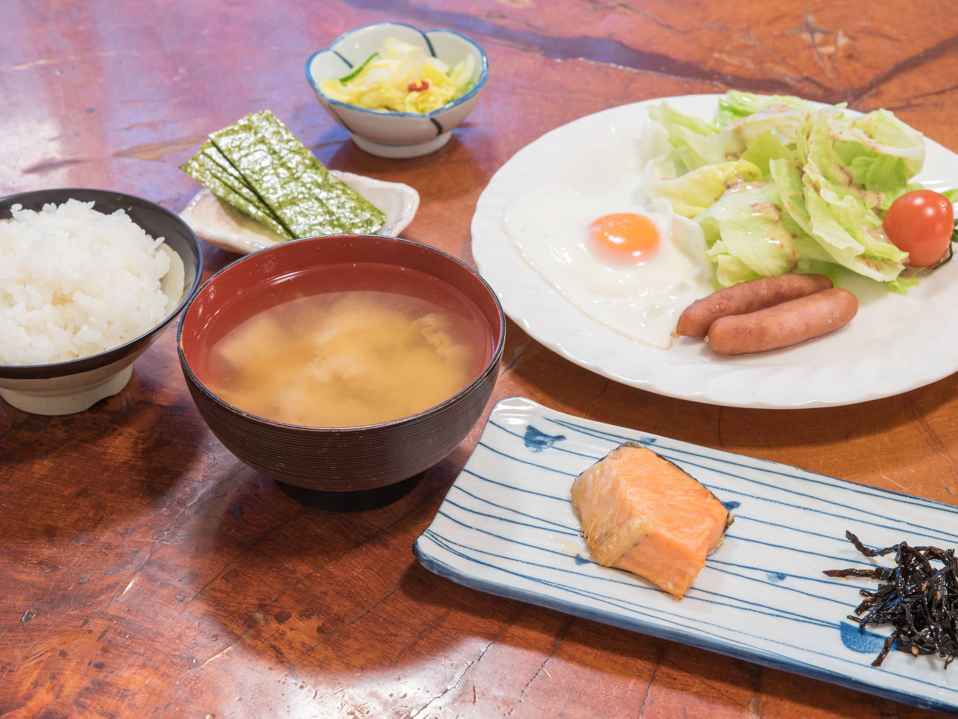 Una mesa con varios platos, incluyendo arroz, sopa, un plato con huevos, lechuga y salchichas, y un plato con salmón y algas.