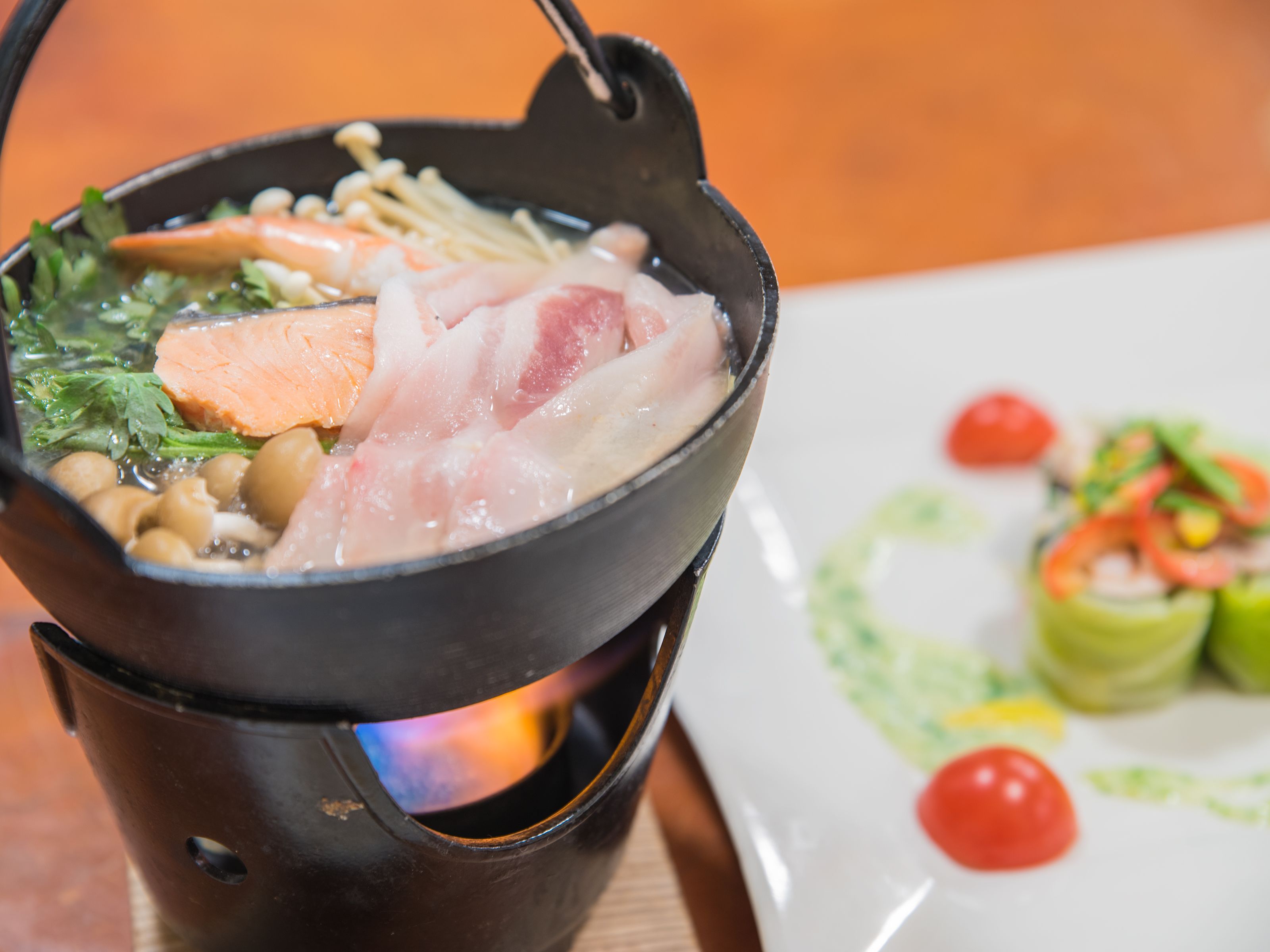 Una olla caliente con diversos ingredientes, un plato de verduras y una ensalada pequeña, sobre una mesa de madera.