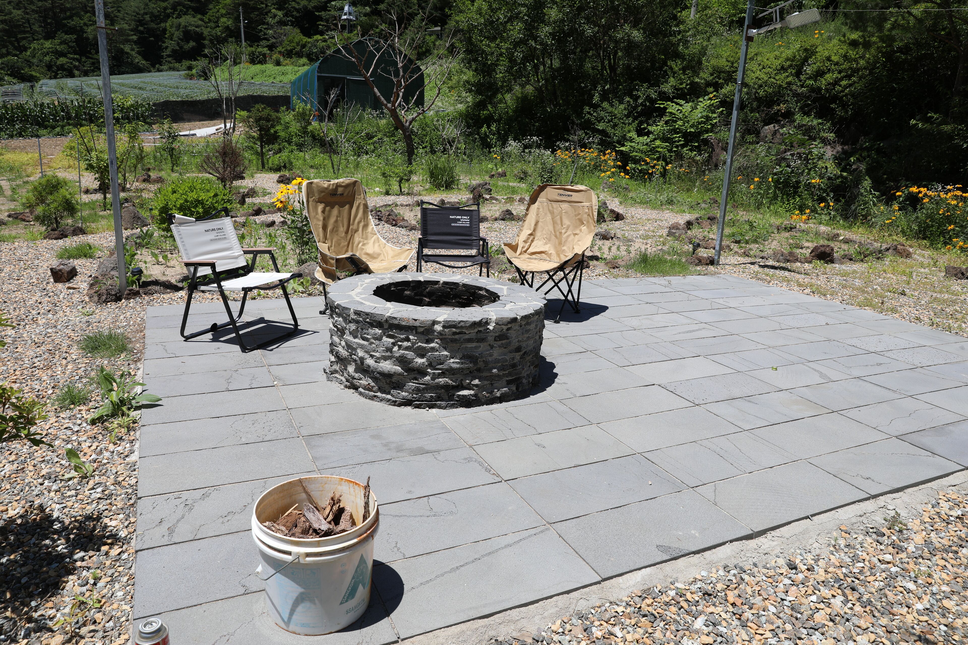 A stone fire pit surrounded by four chairs, a bucket of wood, and a paved patio area.
