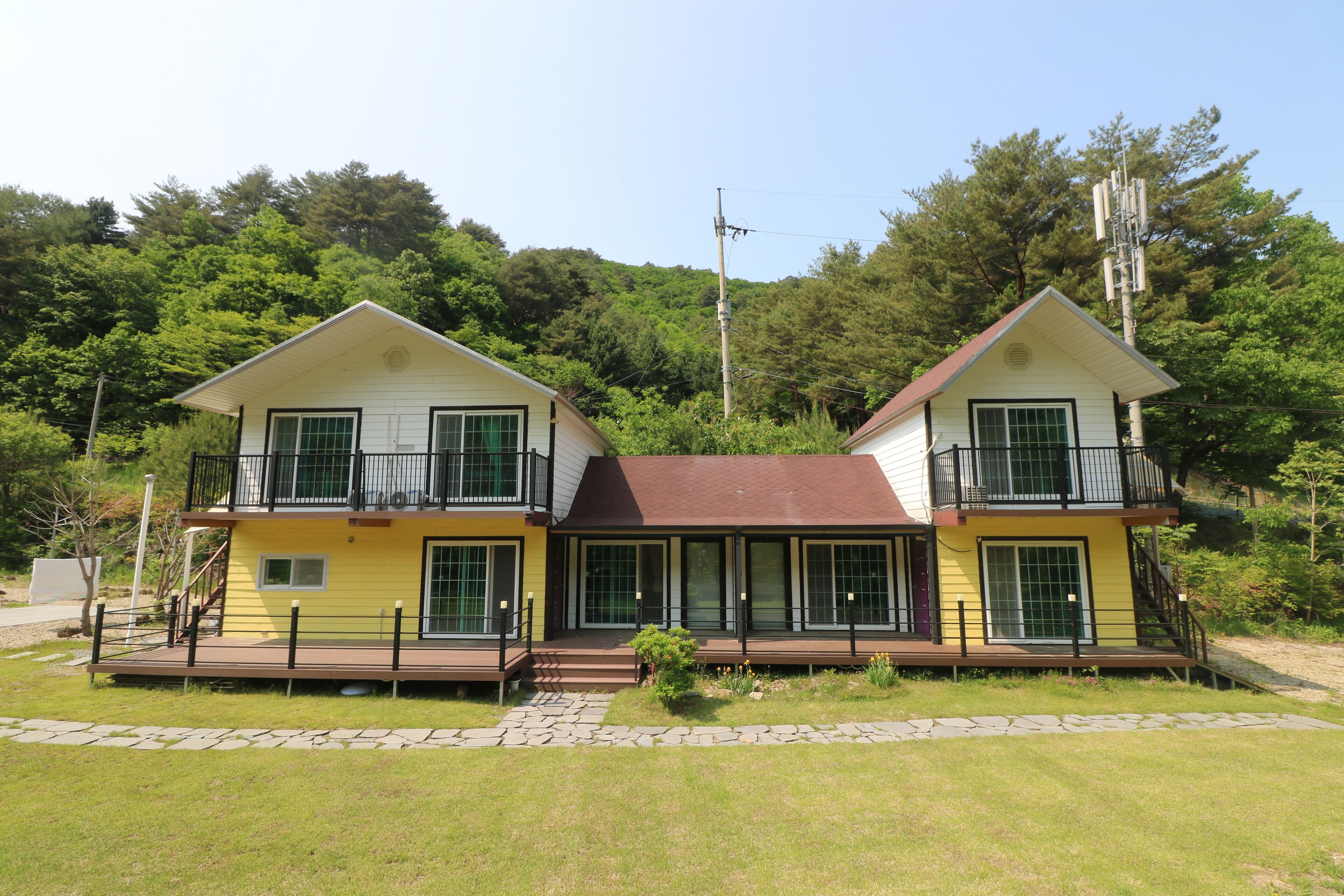 A two-story house with a balcony on each floor, a stone pathway in front, and a forested hillside in the background.