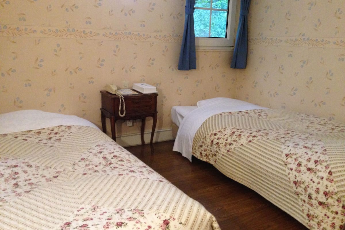 Una habitación con dos camas individuales, una mesita de noche de madera con teléfono y una ventana con cortinas azules.