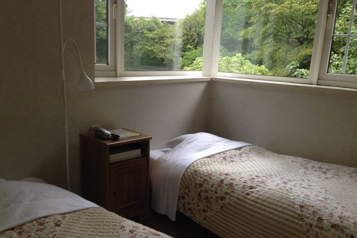 Un dormitorio con dos camas individuales, una mesita de noche, una lámpara y ventana con vistas a la vegetación.