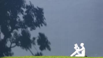 Una escultura blanca de una persona leyendo en un área cubierta de césped, con un árbol de fondo.
