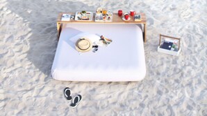 Un coin pique-nique aménagé sur la plage, avec une table, de la nourriture, des boissons et un chapeau posé sur un coussin blanc.