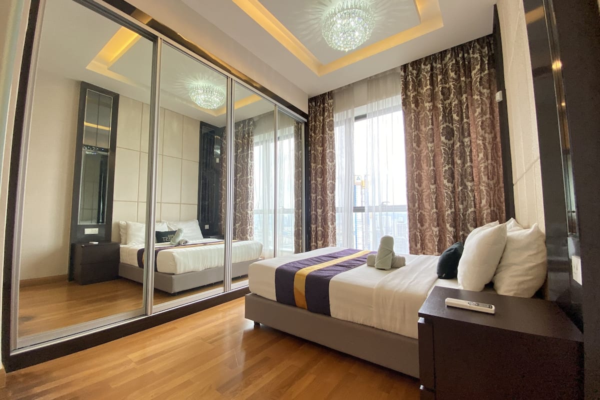 A modern hotel room with a large mirror, a bed with a striped bedspread, a bedside table, and a view of the city through large windows.
