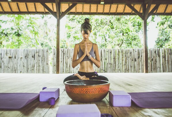 A person meditating in a wooden room with a bamboo fence, a large bowl, and purple mats.