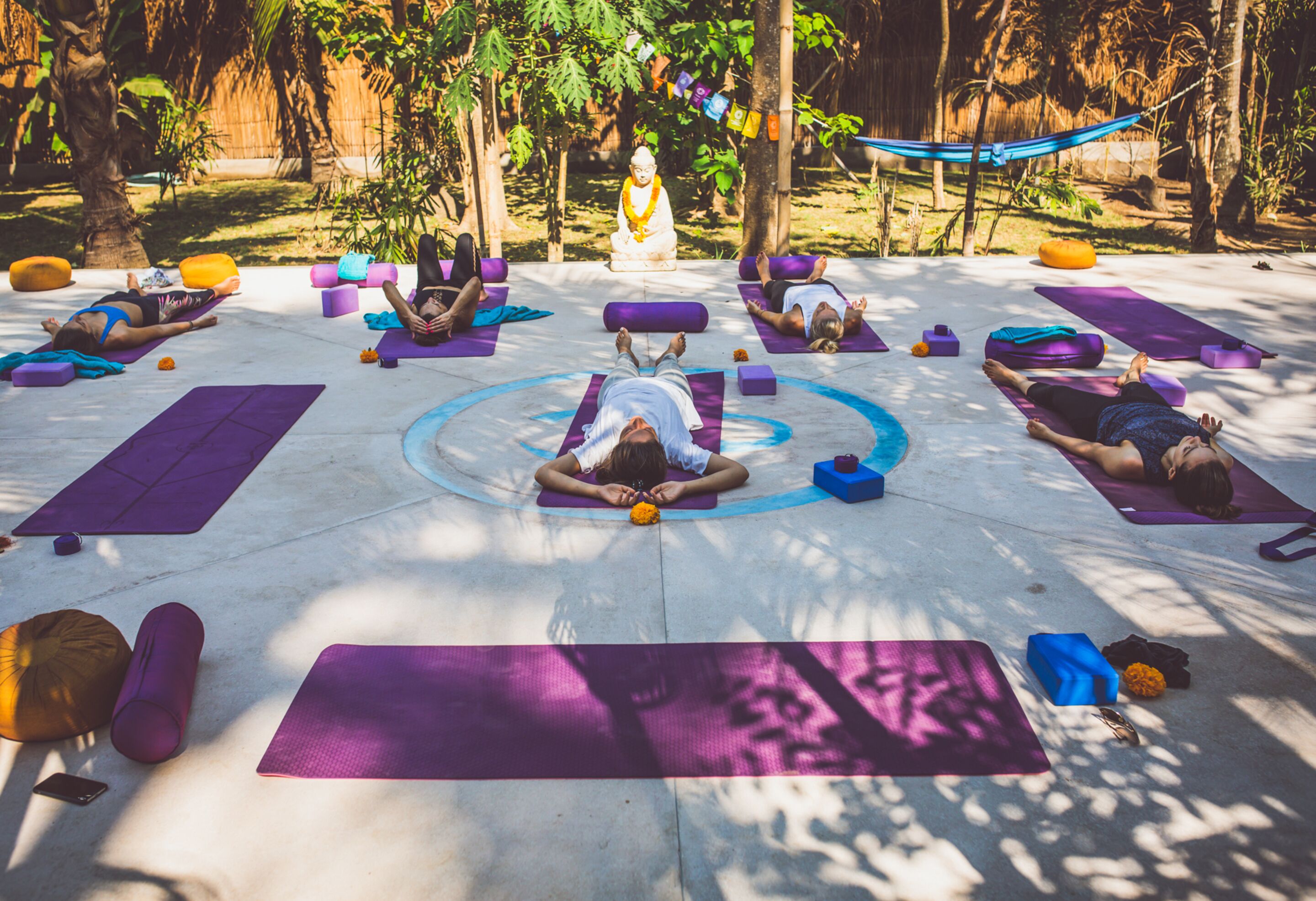 A group of people practicing yoga outdoors on mats arranged in a circle, with a statue and a hammock in the background.