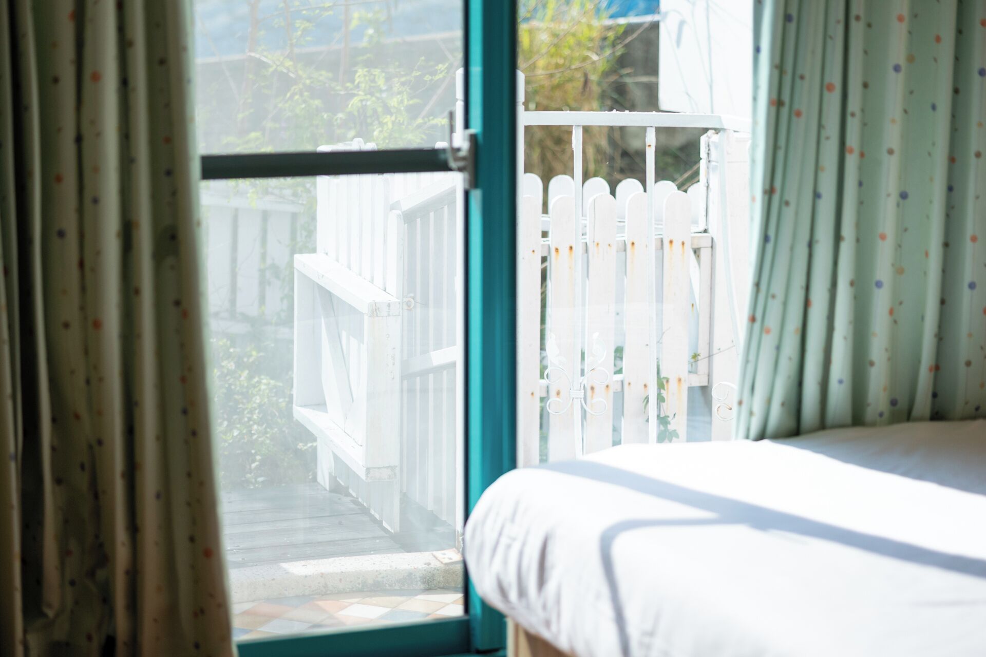 A bedroom with a window, a white bed, and a view of a white picket fence.