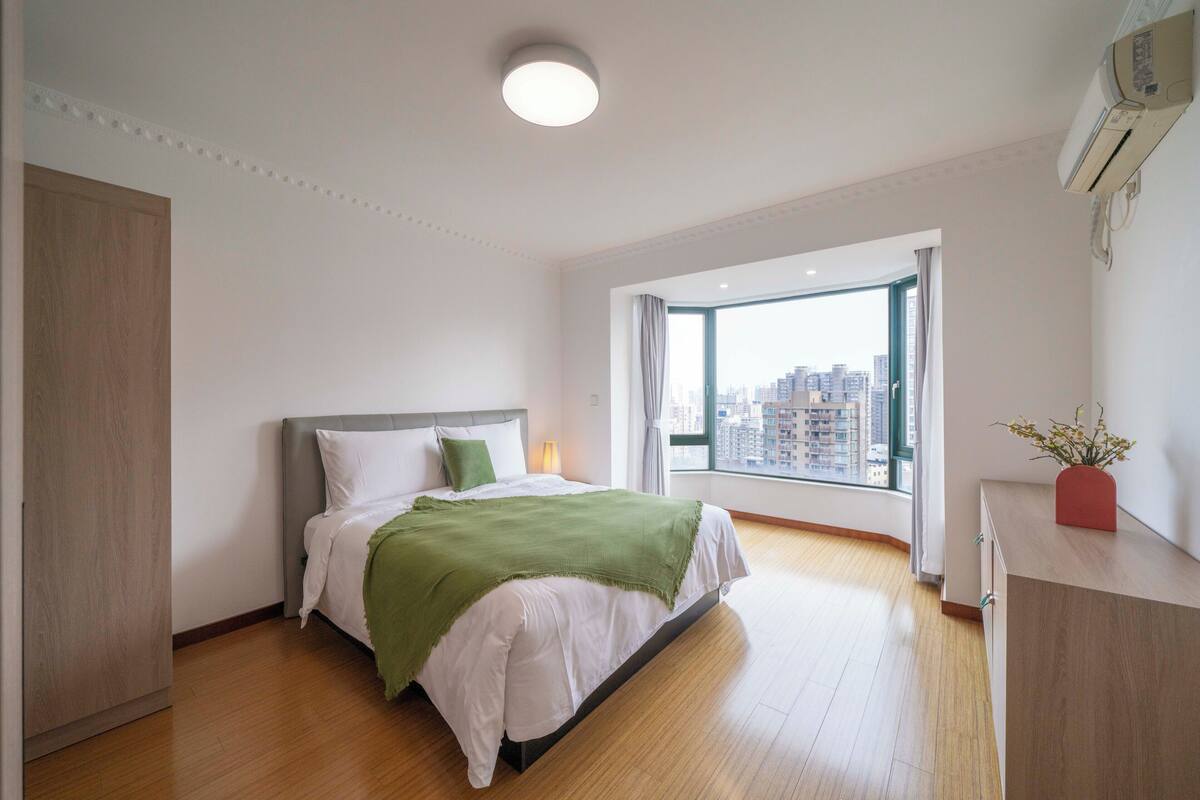 A bedroom with a bed, a window with a city view, a bedside lamp, and a wall-mounted air conditioner.