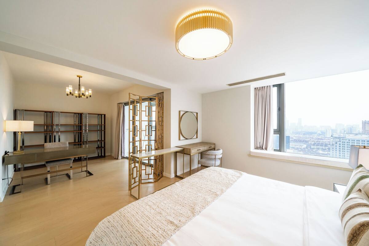 A hotel room with a bed, desk, chair, and a large window with a city view.