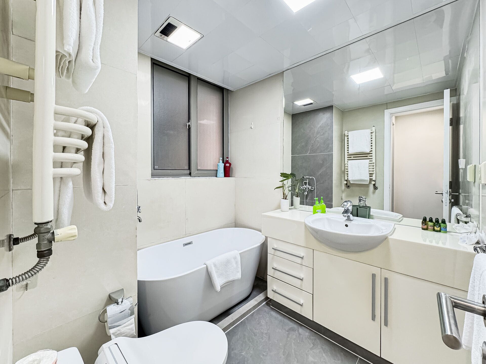 Une salle de bain moderne avec une baignoire, un lavabo et des toilettes. Des serviettes sont accrochées au mur, et il y a une fenêtre au-dessus de la baignoire.