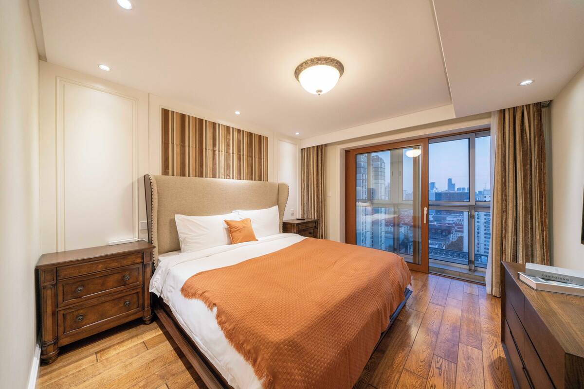 A hotel room with a large bed, a nightstand, a dresser, a window with a city view, and a ceiling light.