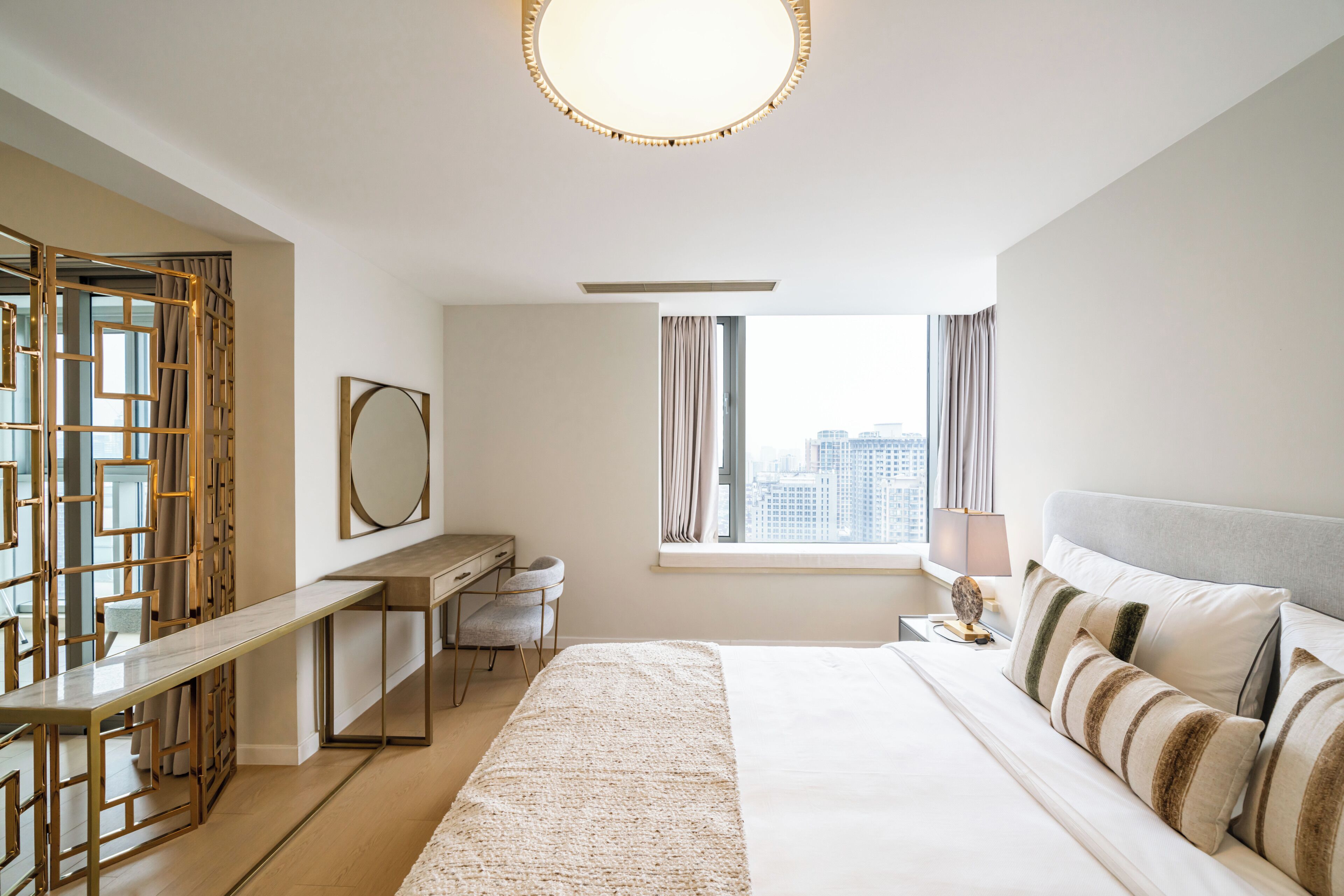 A hotel room with a bed, desk, chair, and a view of the city through the window.