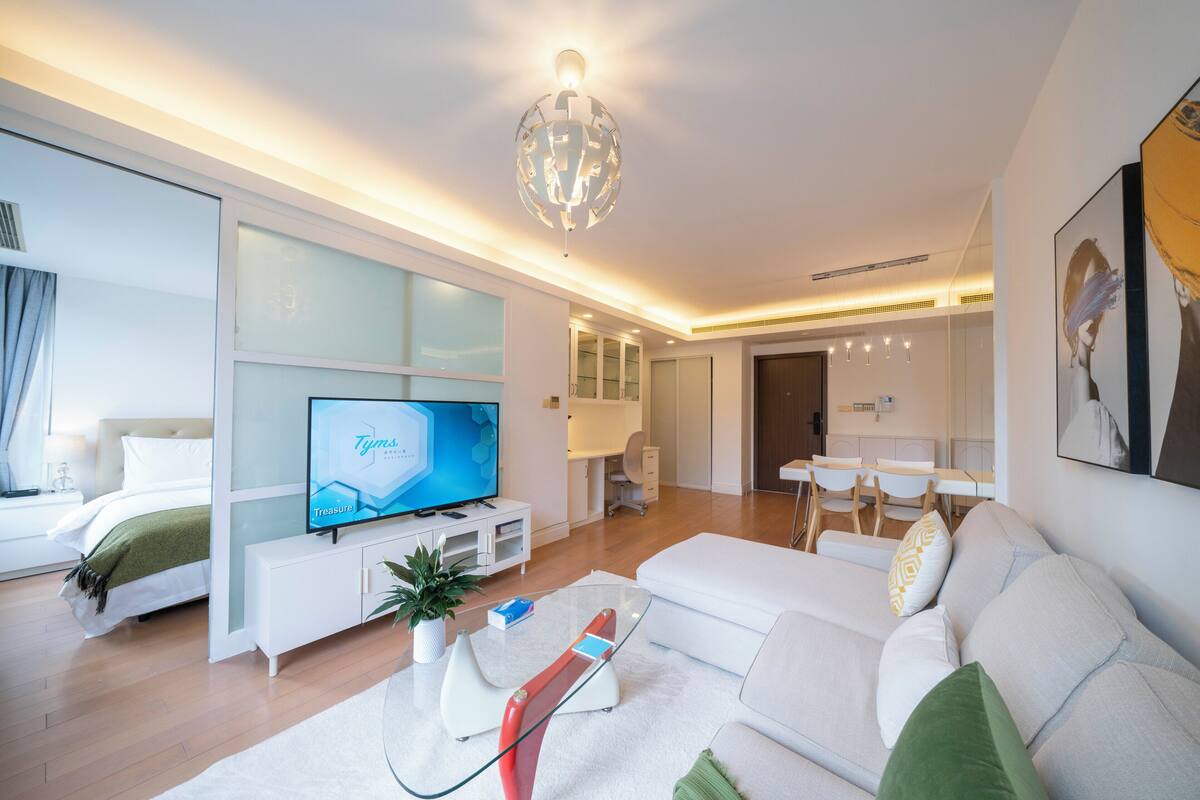 A modern living room with a flat-screen TV, white sofa, glass coffee table, and a dining area with a white table and chairs.