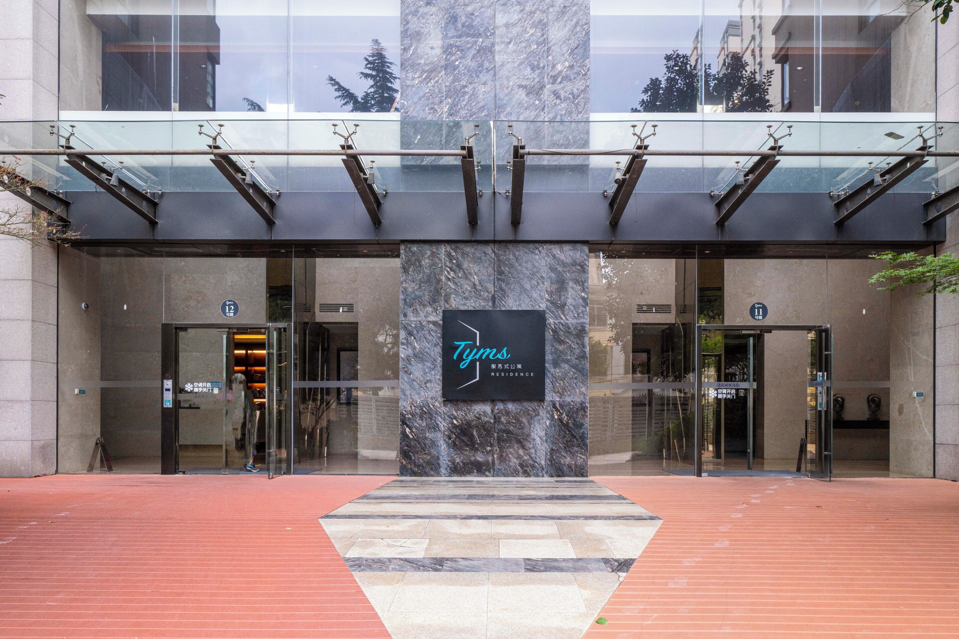 A modern building entrance with a glass facade, a central marble pillar with the name 'Tyns Residence', and a red brick pathway leading to the doors.