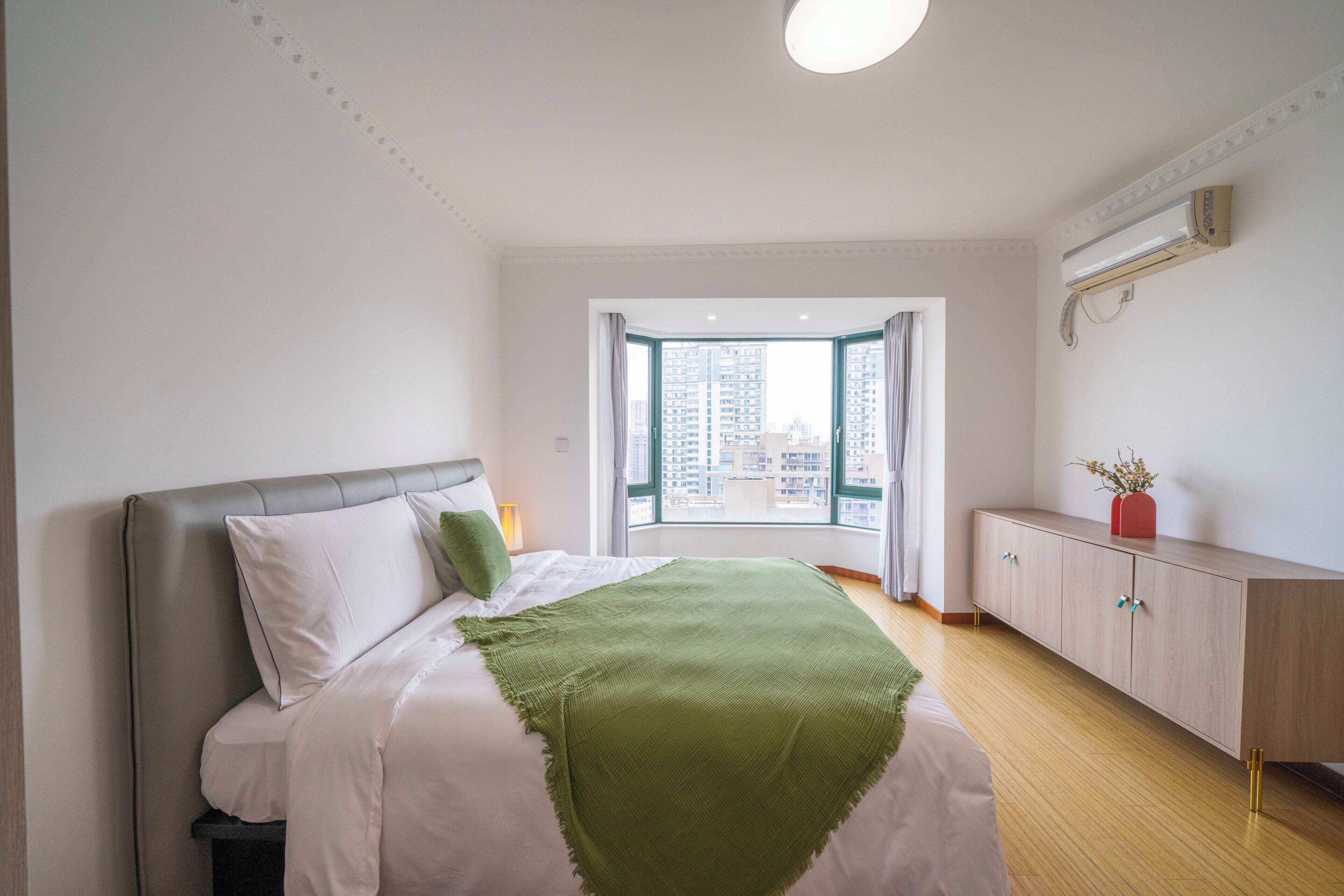 A bedroom with a bed, a nightstand, a dresser, a window with curtains, and a view of buildings.
