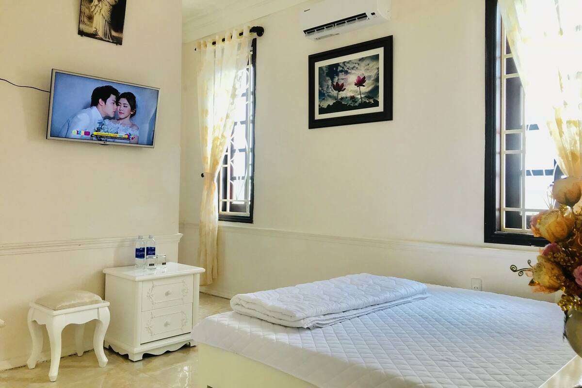 A hotel room with a bed, a TV, a small bedside table, a nightstand, a window with curtains, and a wall-mounted air conditioner.