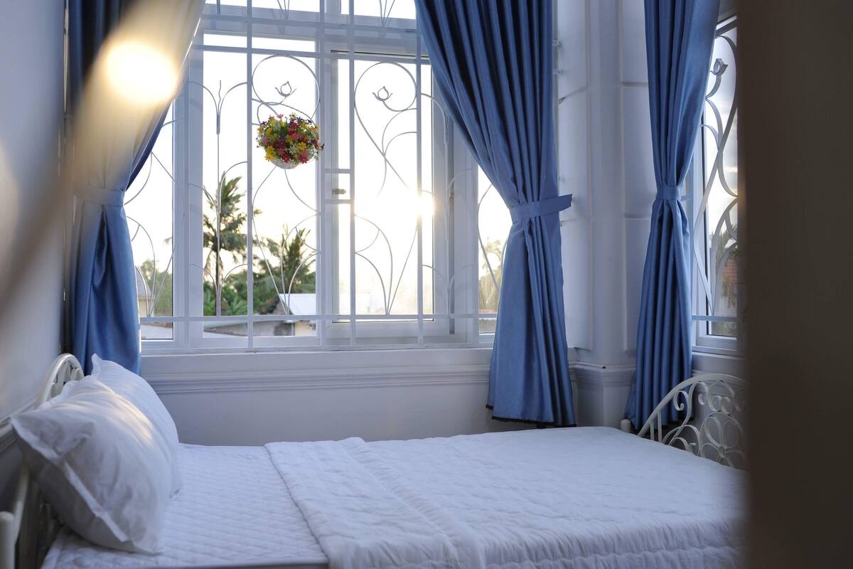 A bedroom with a bed, white curtains, and a window with a view of palm trees.