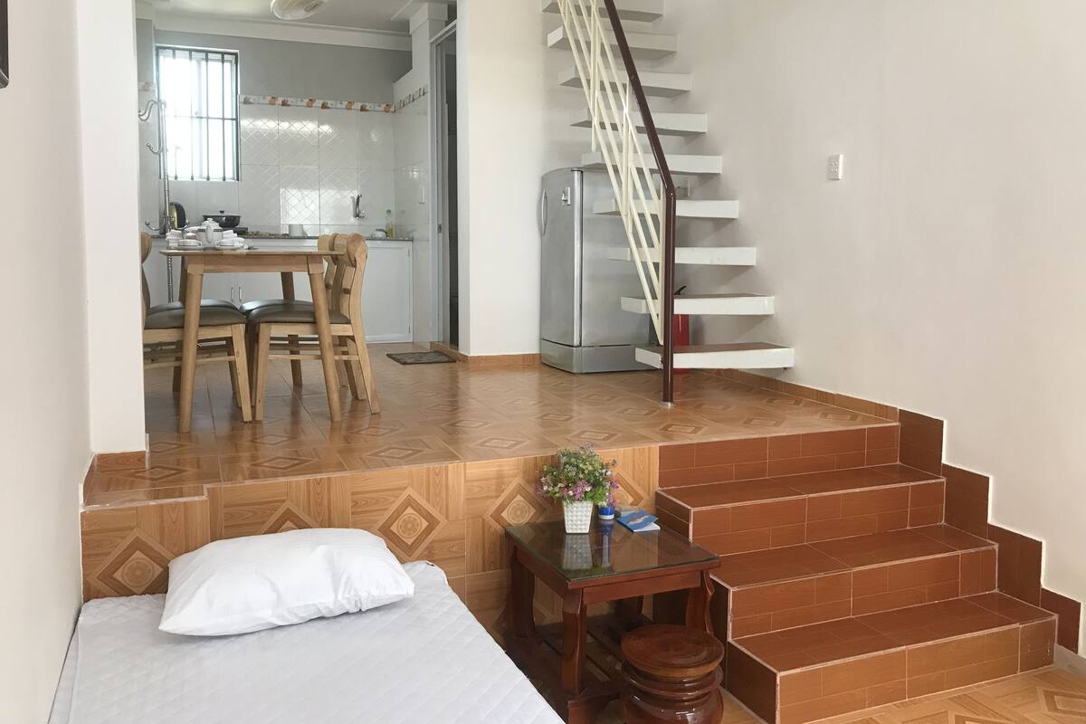 A bedroom with a bed, a wooden headboard, a small table with a plant, and a staircase leading to a kitchen area.