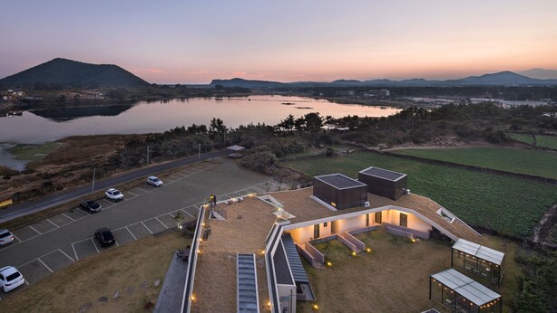 Vista aérea de un complejo moderno de edificios con estacionamiento, mobiliario exterior y vistas a un lago y montañas.