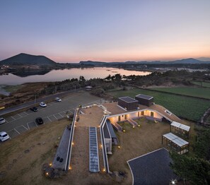 An aerial view of a modern building complex with a parking area, outdoor seating, and a view of a lake and mountains.