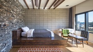 Un dormitorio moderno con pared de ladrillo, cama de madera, silla verde y vista a la ciudad a través de amplias ventanas.