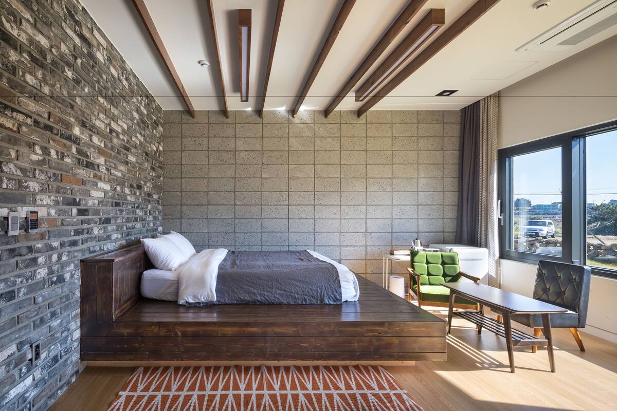 A modern bedroom with a brick wall, a wooden bed, a green chair, and a view of the city through large windows.