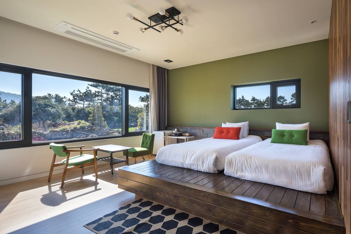 A modern hotel room with a large window, two beds, a desk, and a chair.