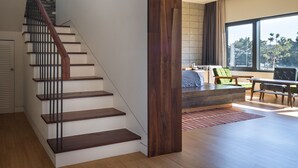 Un interior moderno con una escalera de madera, un gran ventanal que ofrece vistas a árboles y un área de estar con una mesa y sillas.