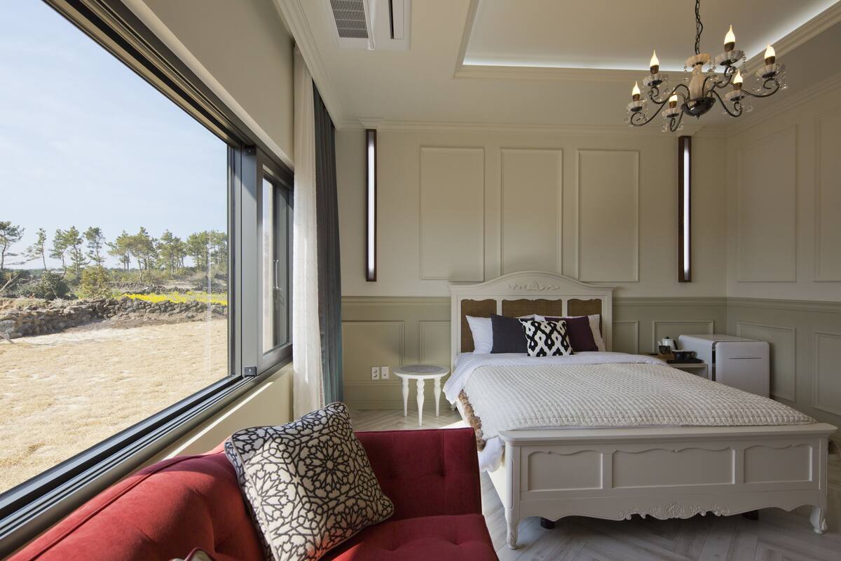 A bedroom with a large window, a bed, a bedside table, and a red chair.