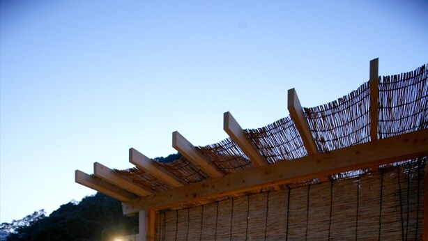 Una terraza de madera con dos sillones de relax y una mesita, una pérgola con techo de tejido, y una zona de hidromasaje.
