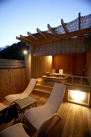 Een houten terras met twee loungestoelen en een kleine tafel, een pergola met een rieten dak, en een zone met een jacuzzi.