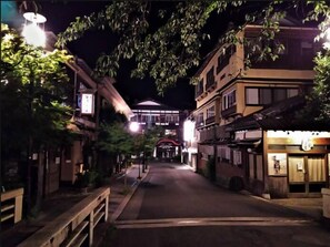 A nighttime street view with buildings on either side, a central road, and a brightly lit building in the distance. - Masutomi Ryokan (Hakone)