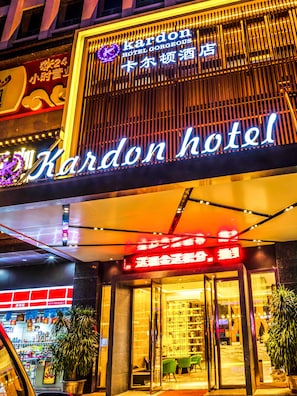 The exterior of Kardon Hotel with a prominent neon sign, a glass entrance, and a well-lit facade.