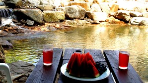 Una mesa de madera con una porción de sandía y dos vasos de bebida roja, frente a un arroyo pedregoso y una cascada.
