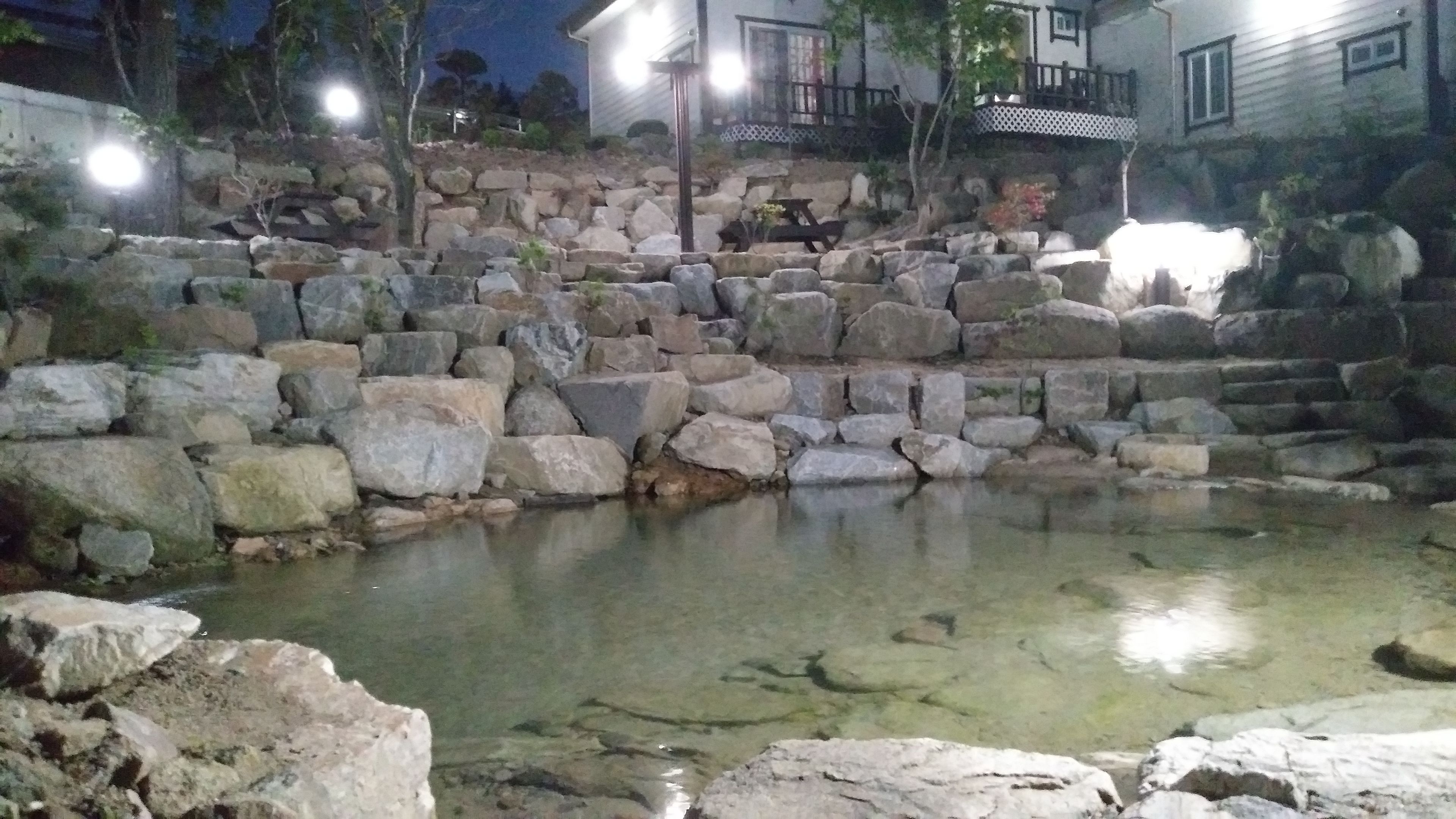 A stone water feature with a clear pool, surrounded by large rocks and a wooden bench.
