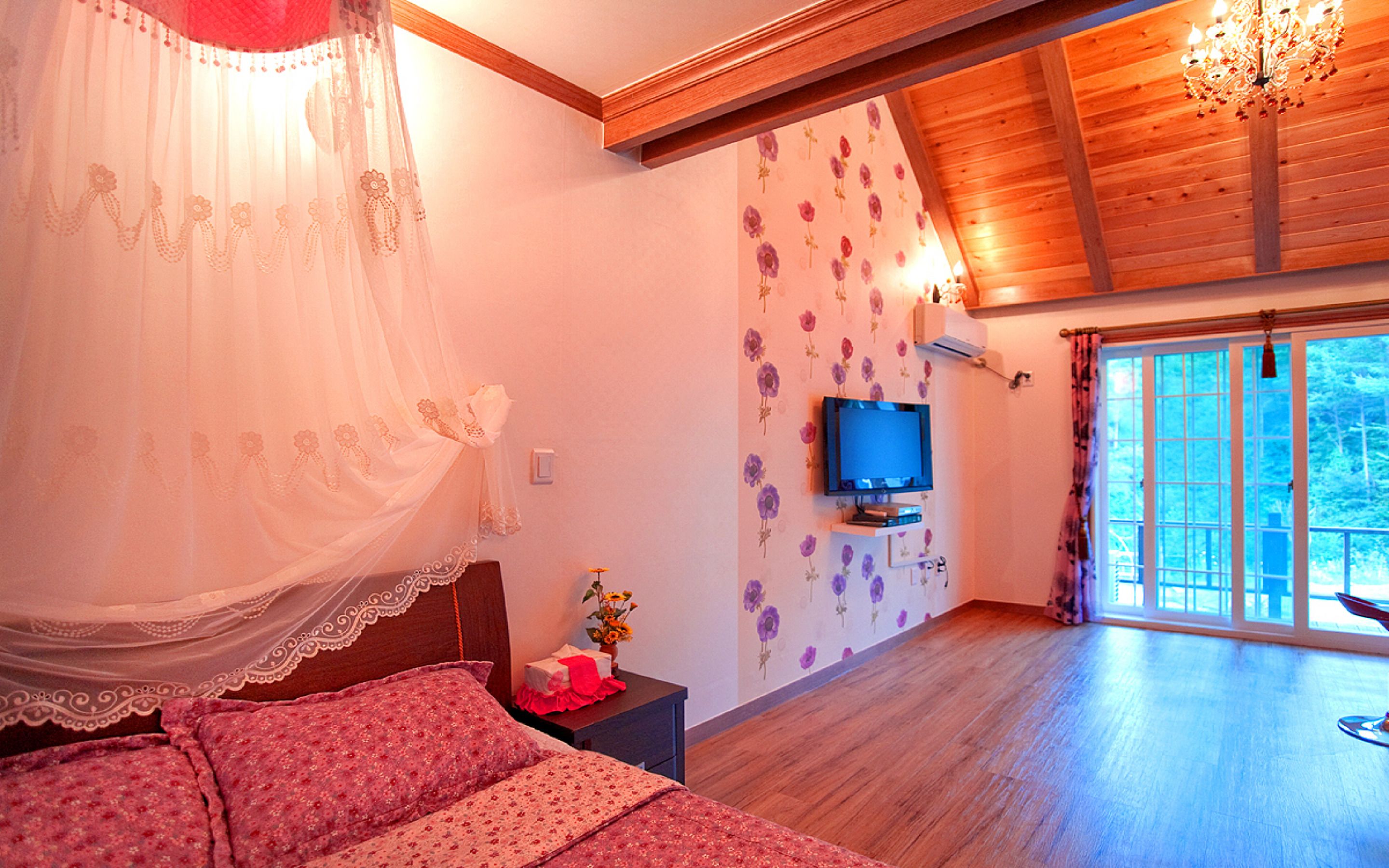 A bedroom with a bed, a TV, a nightstand, a window, and a wooden ceiling.