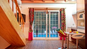 Una cocina con vigas de madera, un comedor con una silla roja, una ventana con cortinas y vista a una piscina.