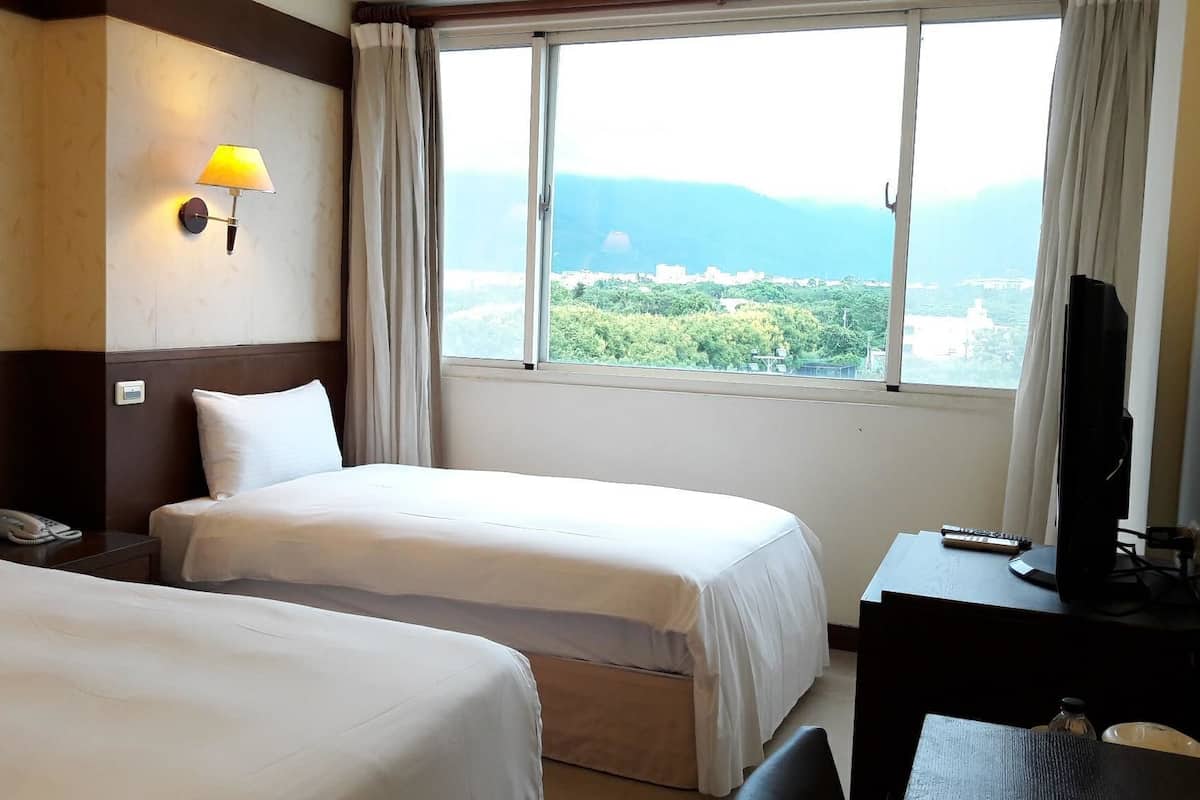 A hotel room with two beds, a desk with a television, a lamp, and a view of a cityscape and mountains.