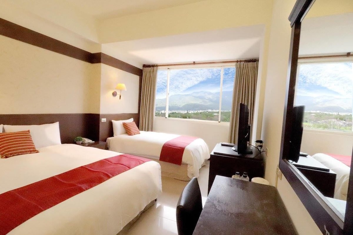 A hotel room with two beds, a desk, a chair, a TV, and a large window with a mountain view.