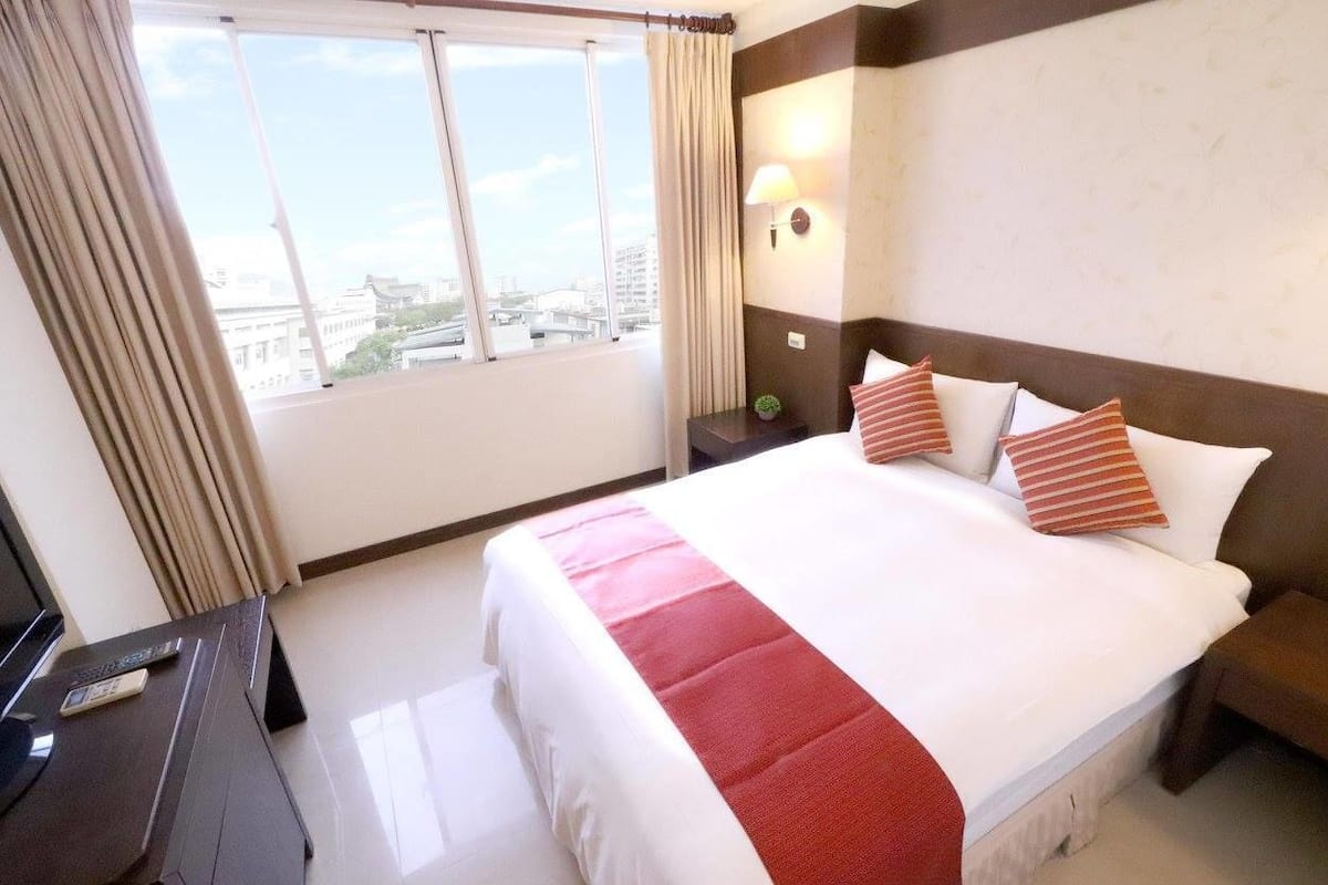 A hotel room with a large bed, a desk with a computer, a lamp, and a view of the city through the window.